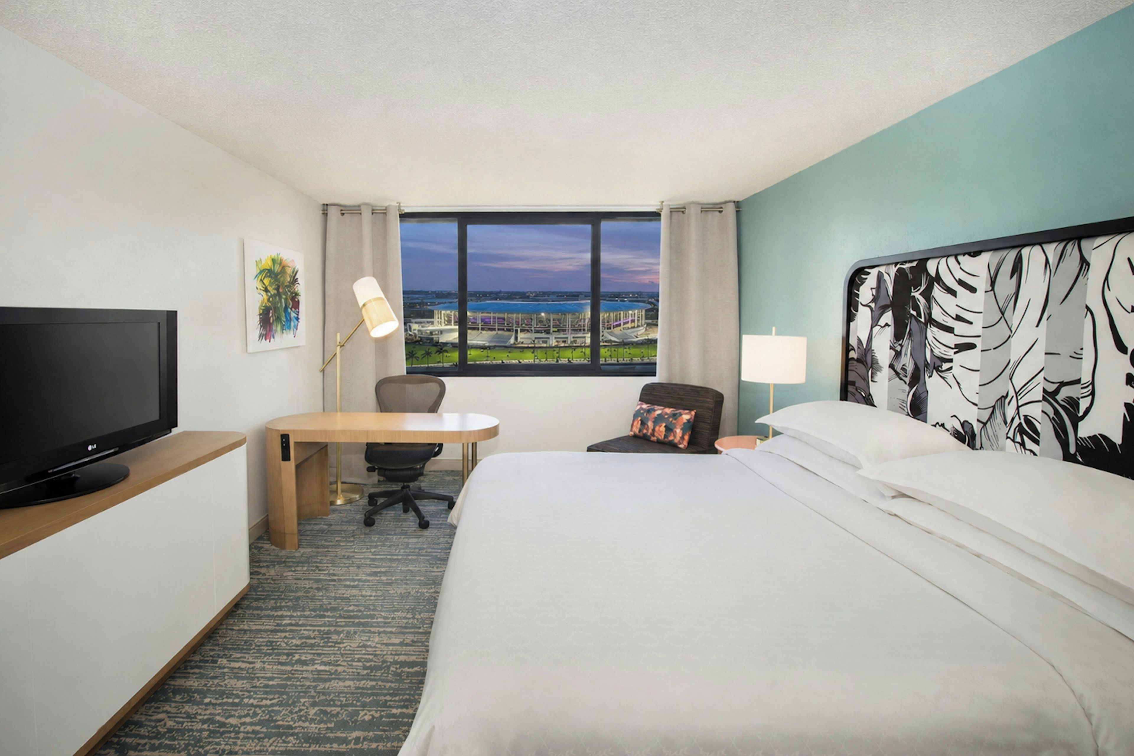 A hotel room with a bed, desk, chair, television, and a window with a view of a distant landscape.