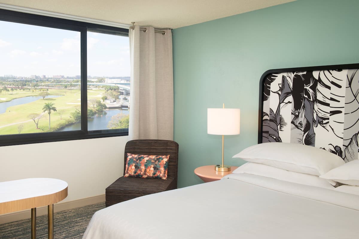 Habitación de hotel con un ventanal grande, una cama con ropa blanca, una mesita redonda y una silla con cojín floral.