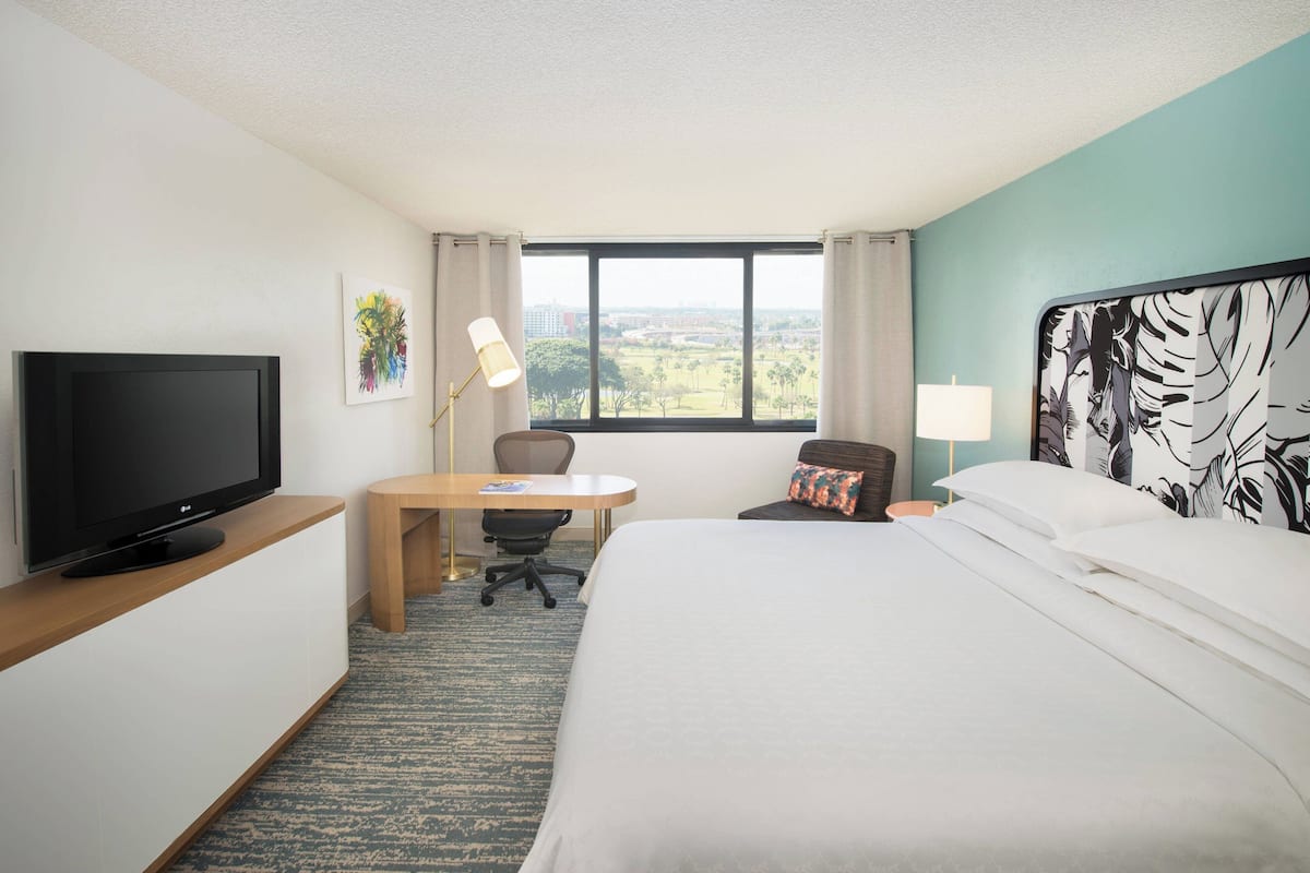 Habitación de hotel con cama, escritorio, televisor, ventana con vista a la ciudad y un cuadro en la pared.