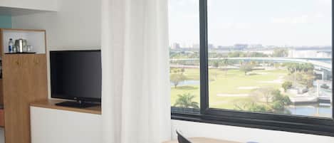 Ein Hotelzimmer mit Fernseher auf einer Holzkommode, einem Schreibtisch mit Stuhl und einem großen Fenster mit Blick auf einen Golfplatz und Stadtpanorama.