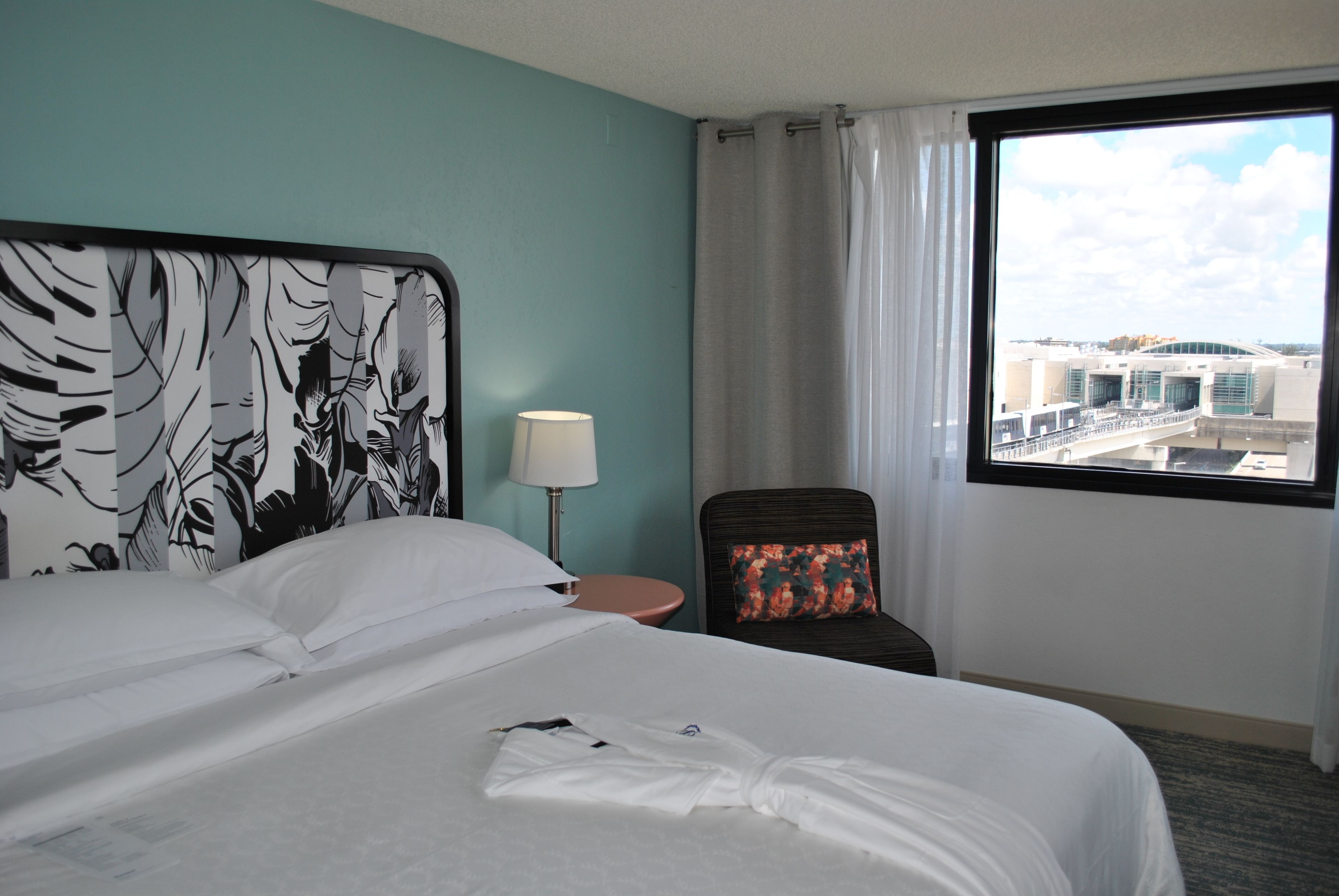 Ein Hotelzimmer mit einem großen Bett, einem Nachttisch, einem Sessel mit einem blumigen Kissen, einer Lampe und einem Fenster mit Blick auf Gebäude.