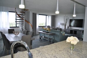 Una habitación de hotel moderna con comedor, sala de estar con sofá y televisor, una escalera y un ventanal grande.