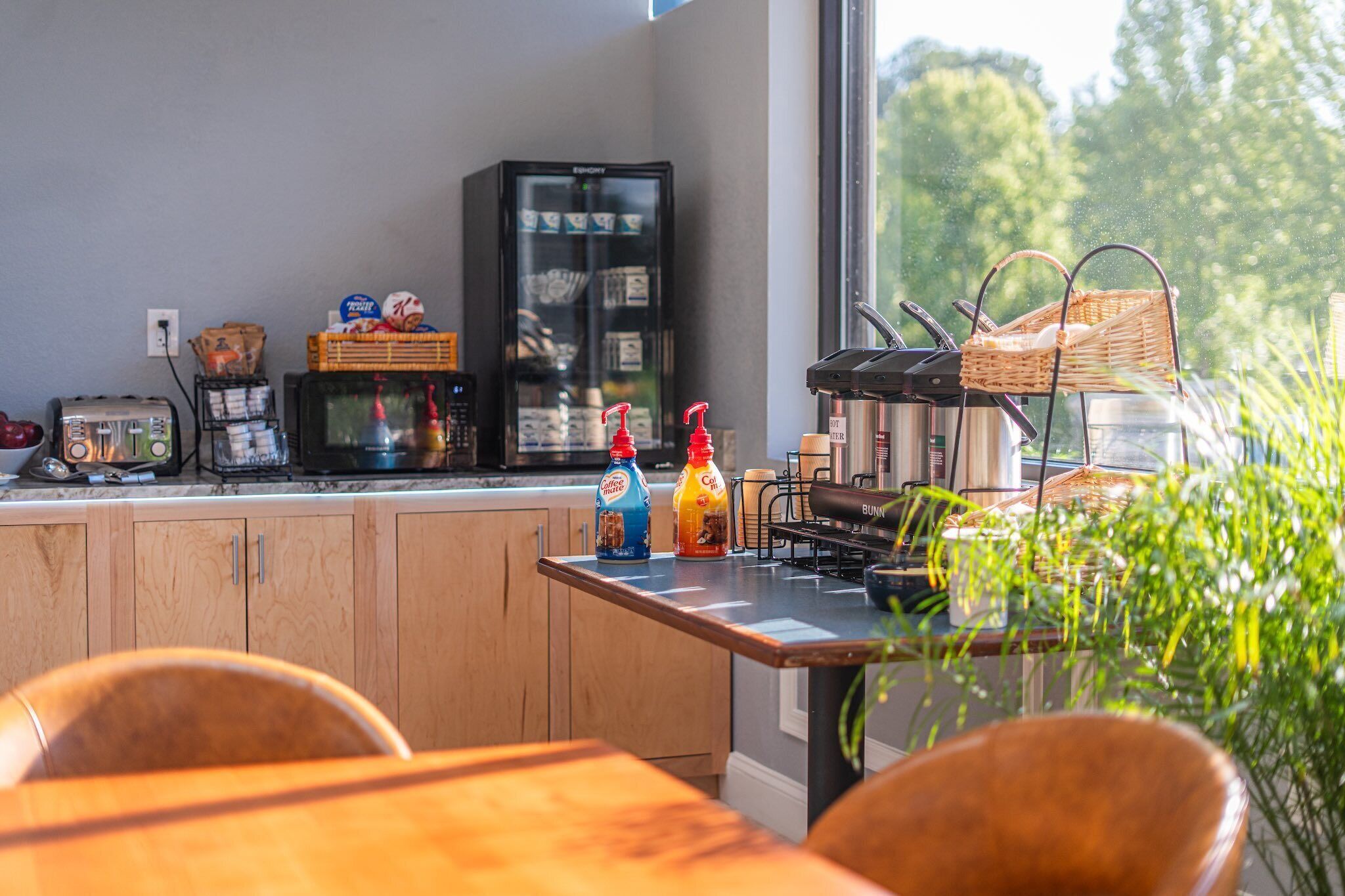 Una cocina equipada con microondas, tostadora, cafetera y dispensador de café. Se observan cestas de mimbre y una ventana con vistas a árboles.