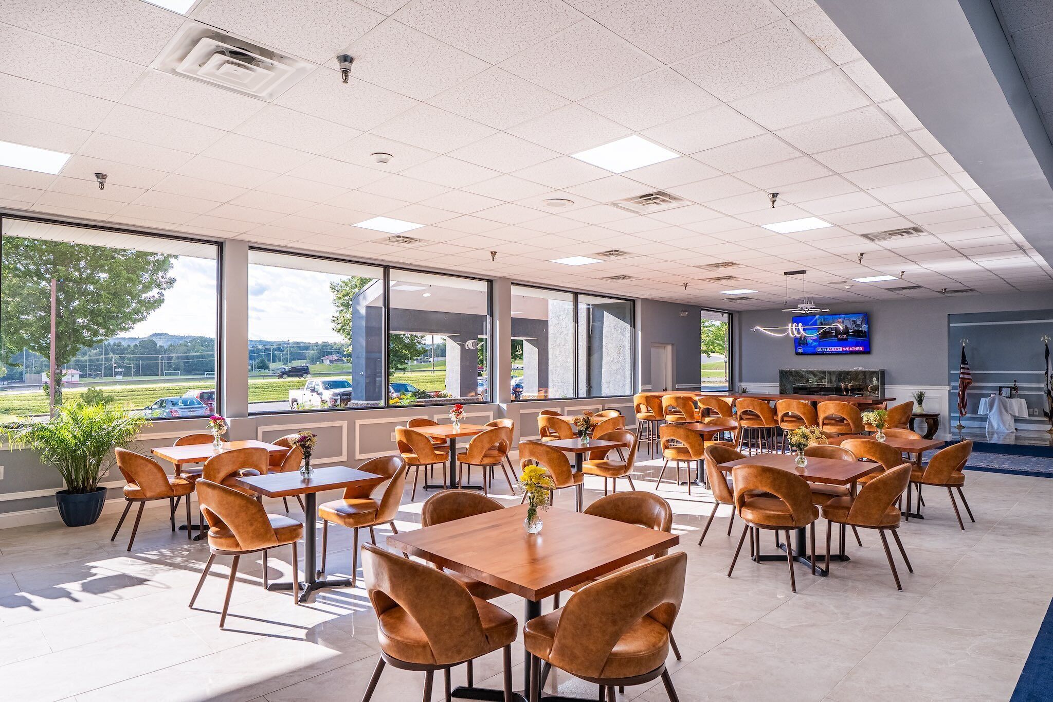 A spacious indoor dining area with multiple tables and chairs, large windows offering a view of the outdoors, and a television mounted on the wall.
