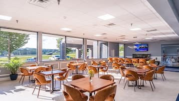 A spacious indoor dining area with multiple tables and chairs, large windows offering a view of the outdoors, and a television mounted on the wall.