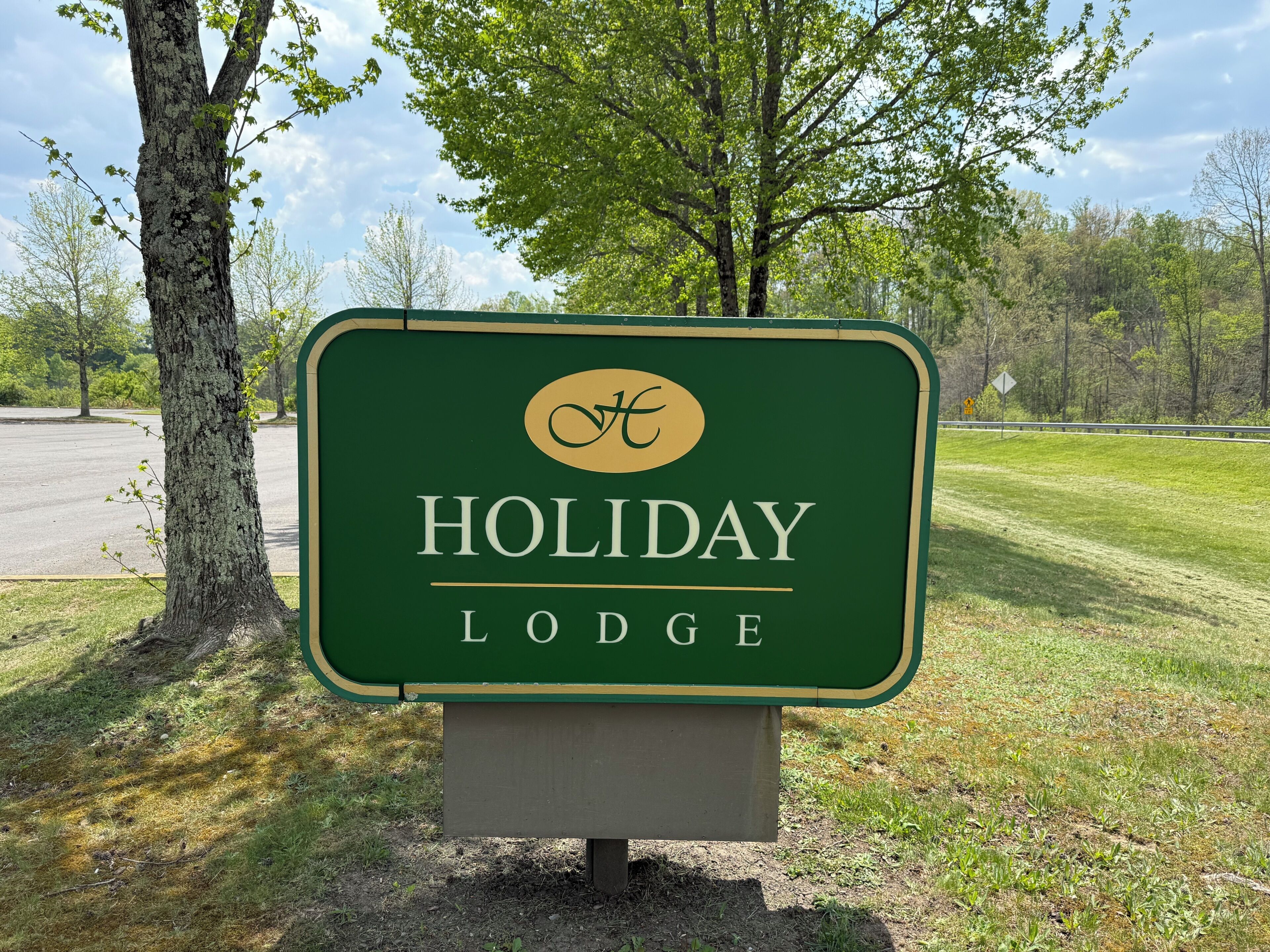 Un panneau vert avec l’inscription « HOLIDAY LODGE » et un logo, le tout sur un fond de forêt et d’un ciel dégagé.