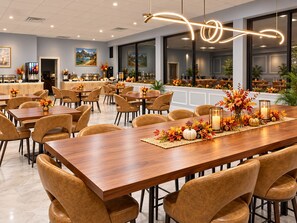 A spacious dining area with multiple tables and chairs, a long wooden table with a centerpiece, and a kitchen area in the background.