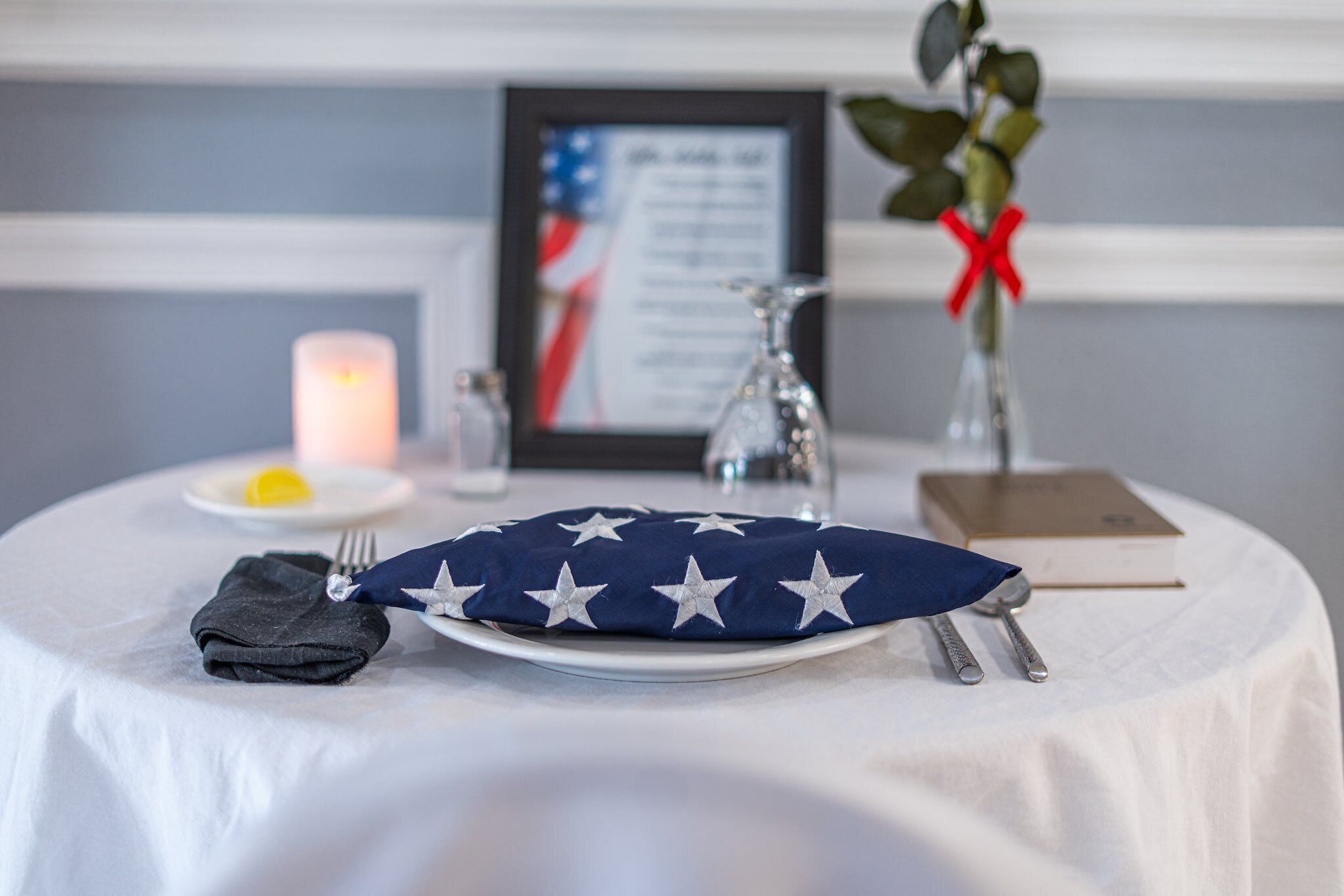 A table with a white tablecloth, a folded dark cloth, a blue pillow with white stars, a framed document, a candle, a salt shaker, a glass, and a red ribbon on a plant.