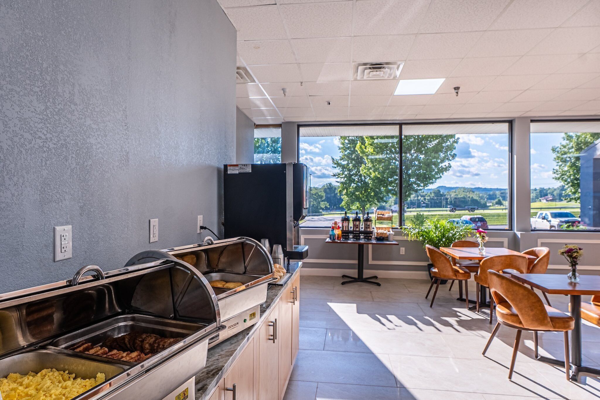A buffet area with various food stations, a seating area with tables and chairs, and a view of the outdoors through large windows.
