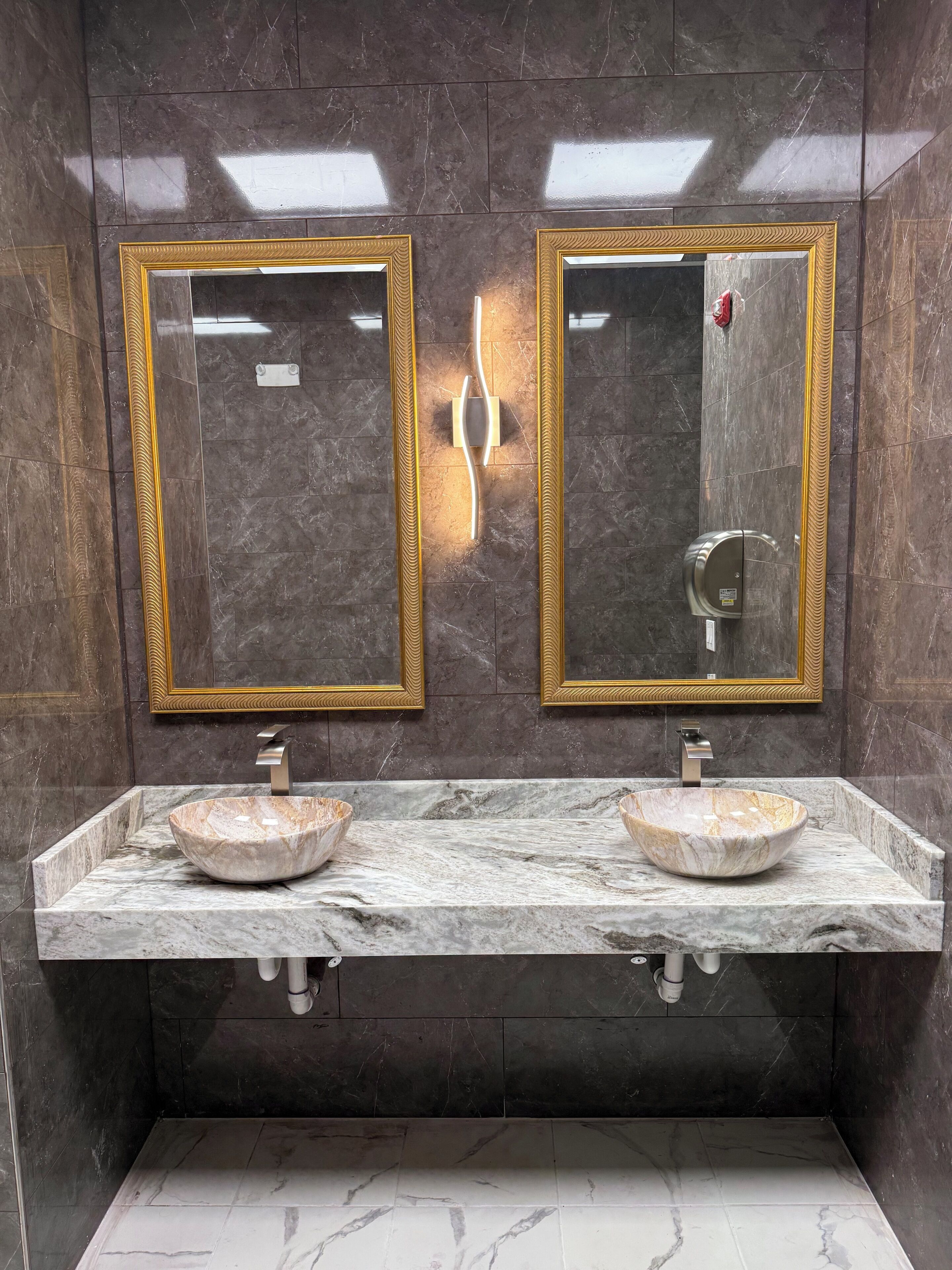 Un baño con dos lavamanos de mármol, espejos con marco dorado y una lámpara de pared.
