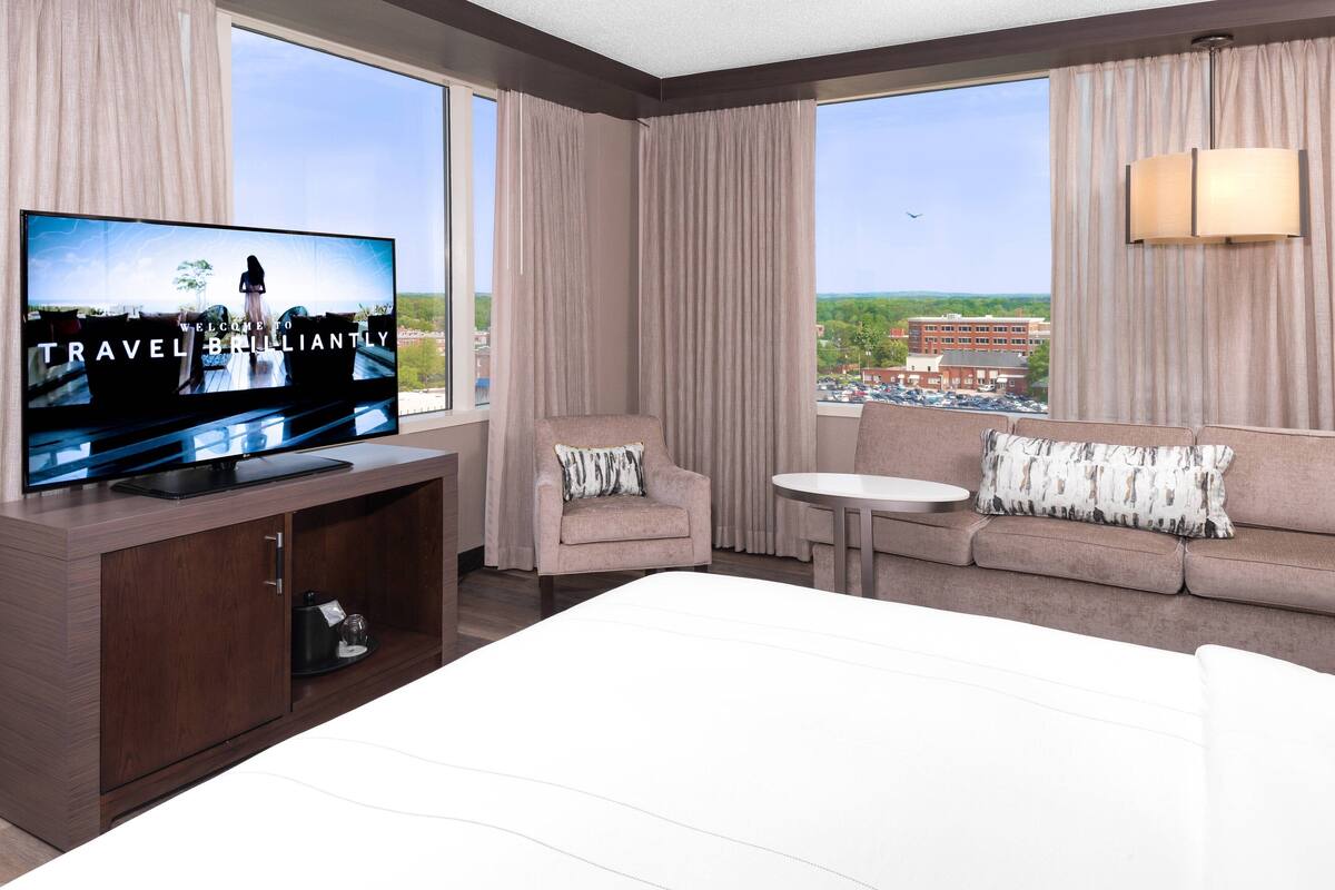 Habitación de hotel con un televisor de pantalla plana grande, un sofá, una silla, una mesita y vistas a la ciudad a través de amplias ventanas.