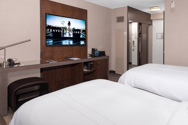 A hotel room with a TV displaying a travel ad, a desk with a phone and a coffee maker, and two beds.