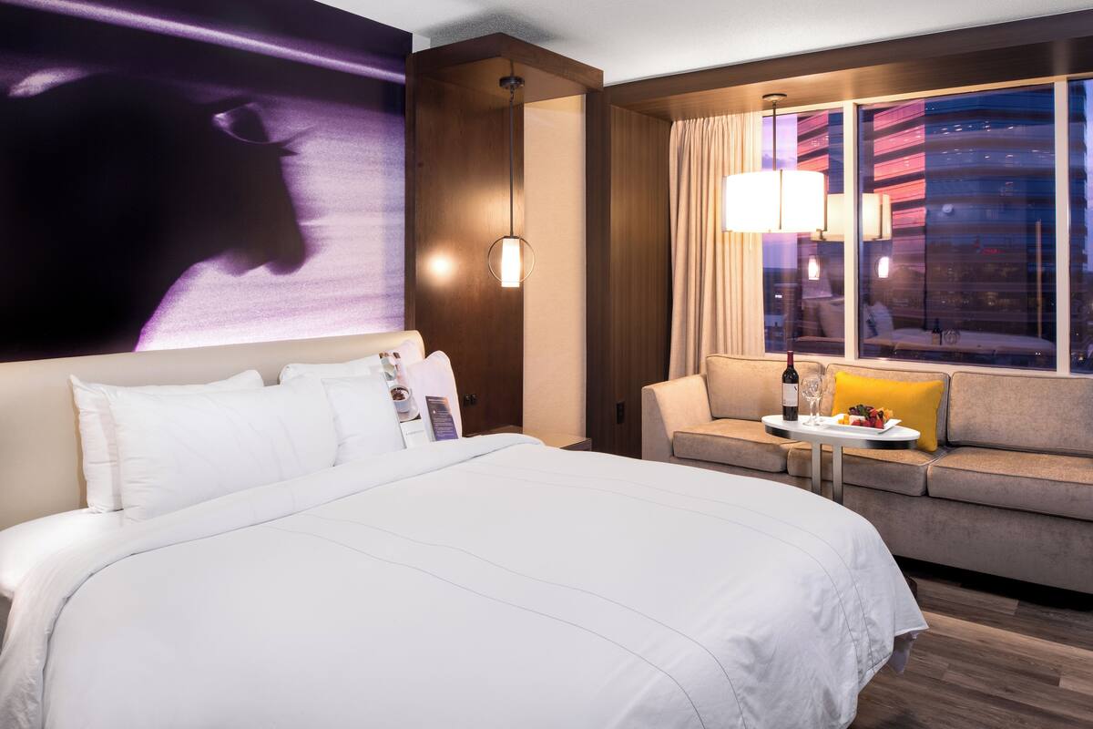 Habitación de hotel con una cama grande, un sofá, una mesita con una botella y vasos, y un ventanal con cortinas.