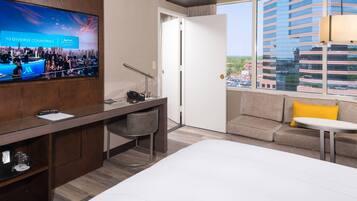 Habitación de hotel con cama, escritorio con silla, televisor, sofá y ventana con vista a la ciudad.