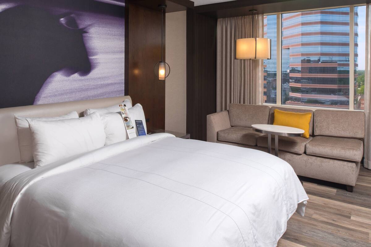 Habitación de hotel con una cama grande, un sofá, una mesita y vistas de la ciudad a través de amplias ventanas.