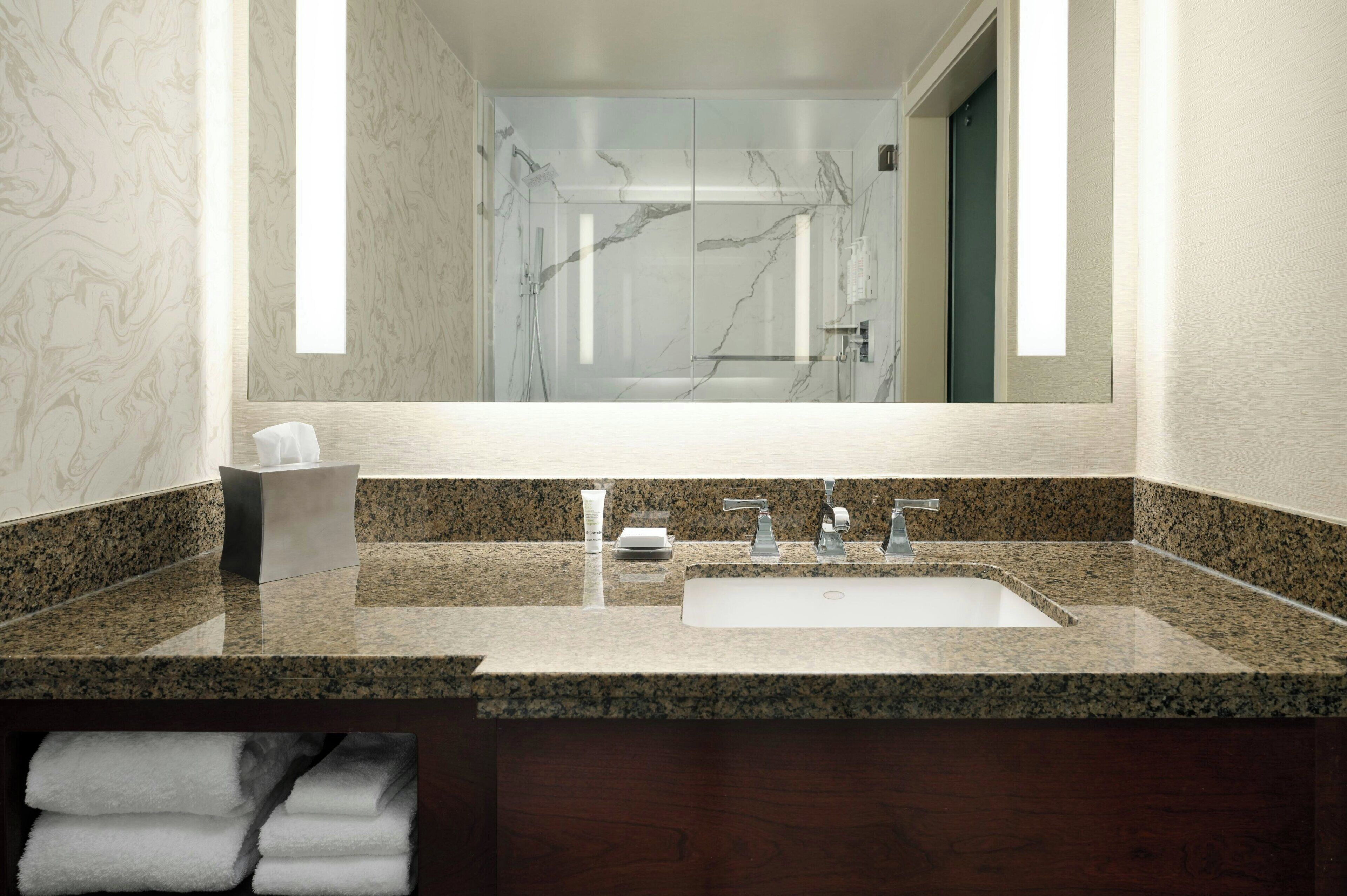 Une salle de bain avec un comptoir en granit, un lavabo, un miroir et une douche dont on aperçoit le reflet.