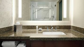 Une salle de bain avec un comptoir en granit, un lavabo, un miroir et une douche dont on aperçoit le reflet.