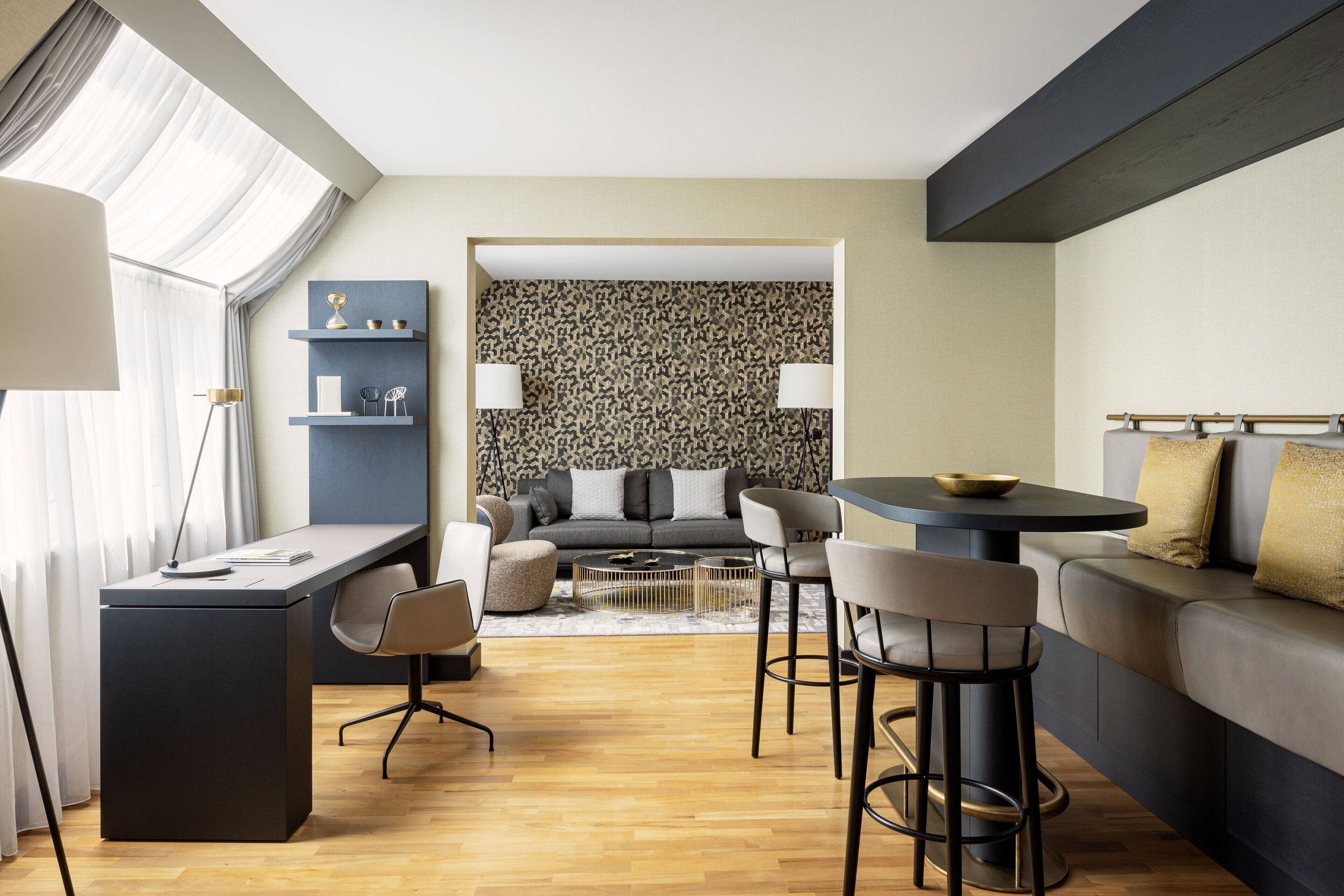 A modern living room with a sofa, a dining table, and a desk. There is a patterned wallpaper on the back wall and a window with curtains.