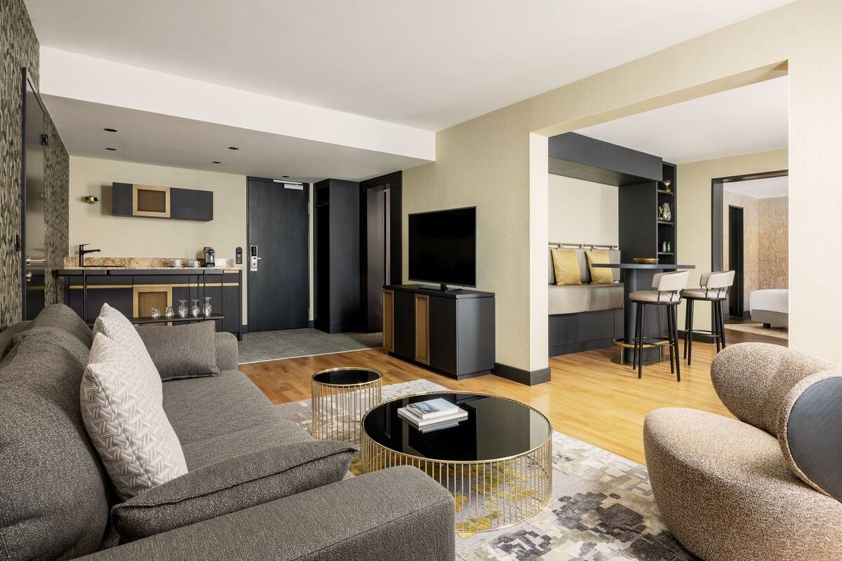 A modern living room with a sofa, coffee table, and a TV. There is a kitchen area with a microwave and cabinets.