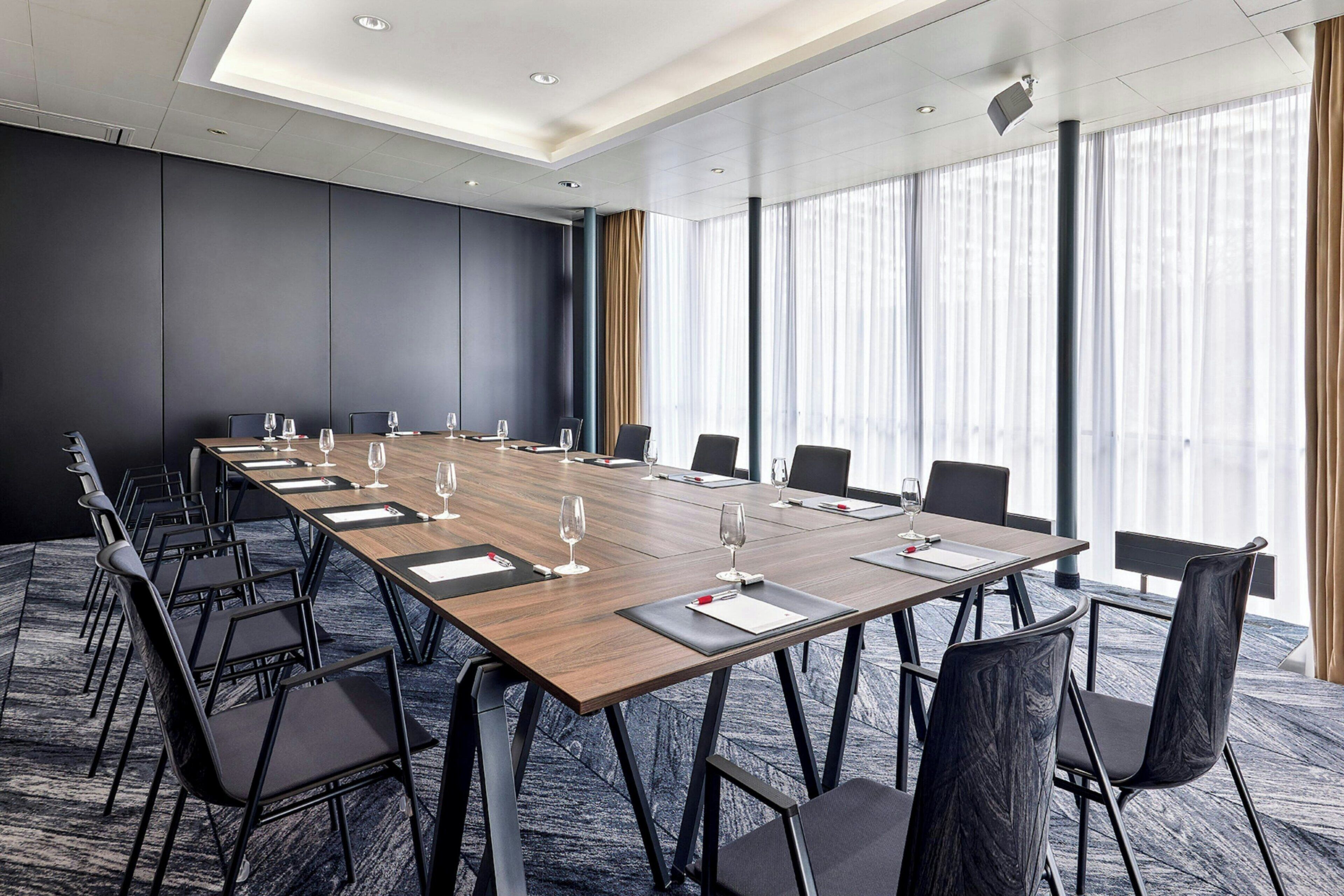 Un salón de conferencias con una larga mesa, sillas negras y ventanas amplias con cortinas.