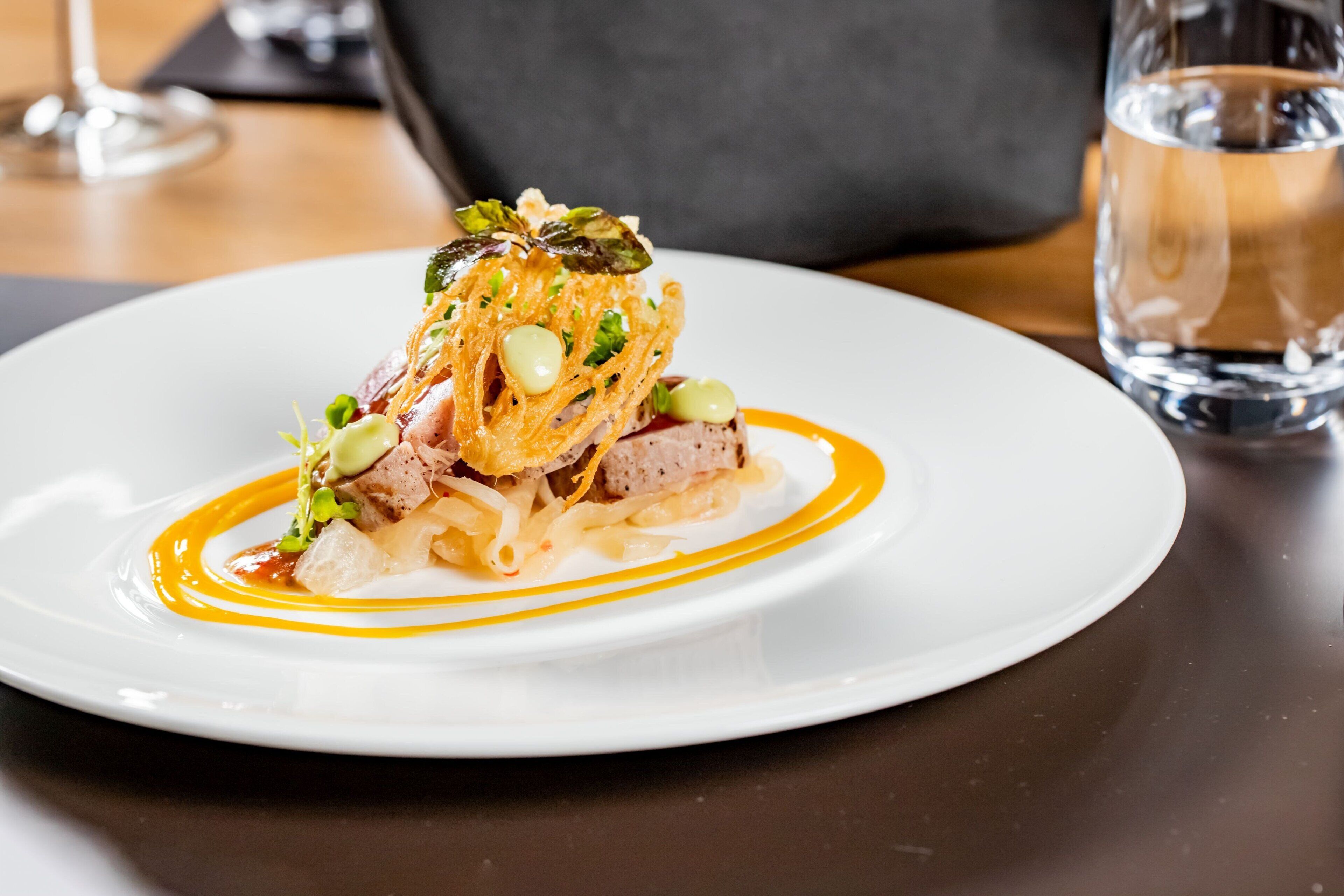 A white plate with a gourmet dish, a glass of water, and a dark-colored napkin on a wooden table.