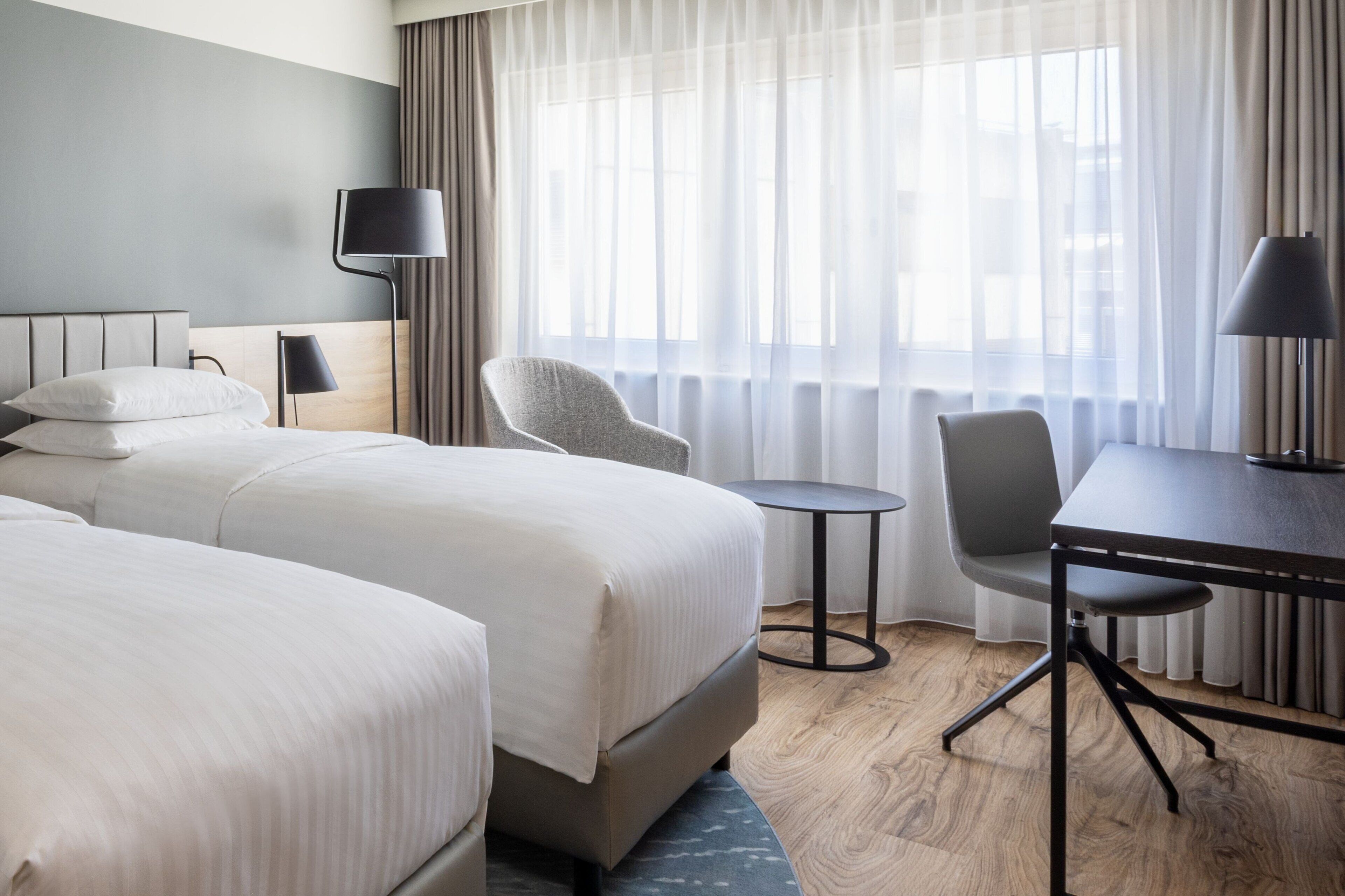 Ein Hotelzimmer mit zwei Betten, einem kleinen runden Tisch, zwei Stühlen, einem Schreibtisch und einer Lampe.
