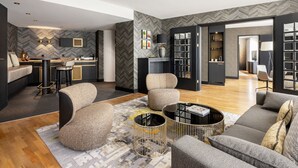 Una sala de estar moderna con un sofá, sillones y una mesa de centro. Se observa una zona de cocina al fondo con una barra y asientos.