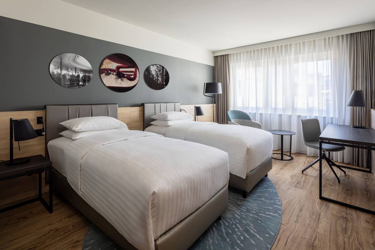 A hotel room with two beds, a desk, a chair, and a lamp.