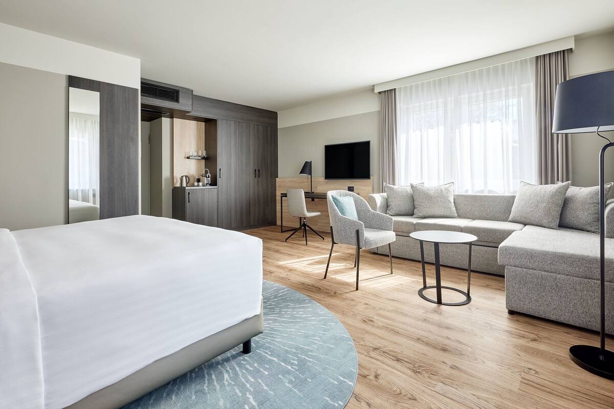 A modern hotel room with a bed, a sofa, a small table, a TV, and a desk.