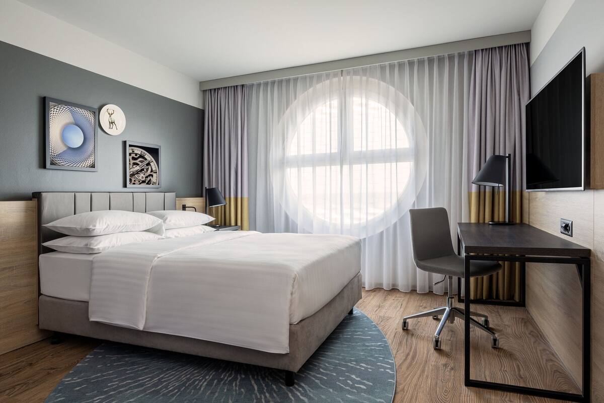 A modern hotel room with a bed, a desk, and a large window with curtains.