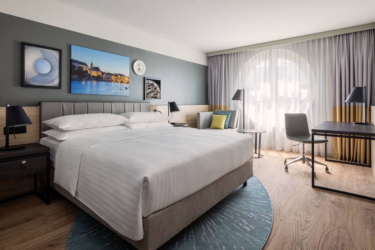 A modern hotel room with a large bed, a desk, and a chair. There are artworks on the walls, a clock, and a view of a cityscape.