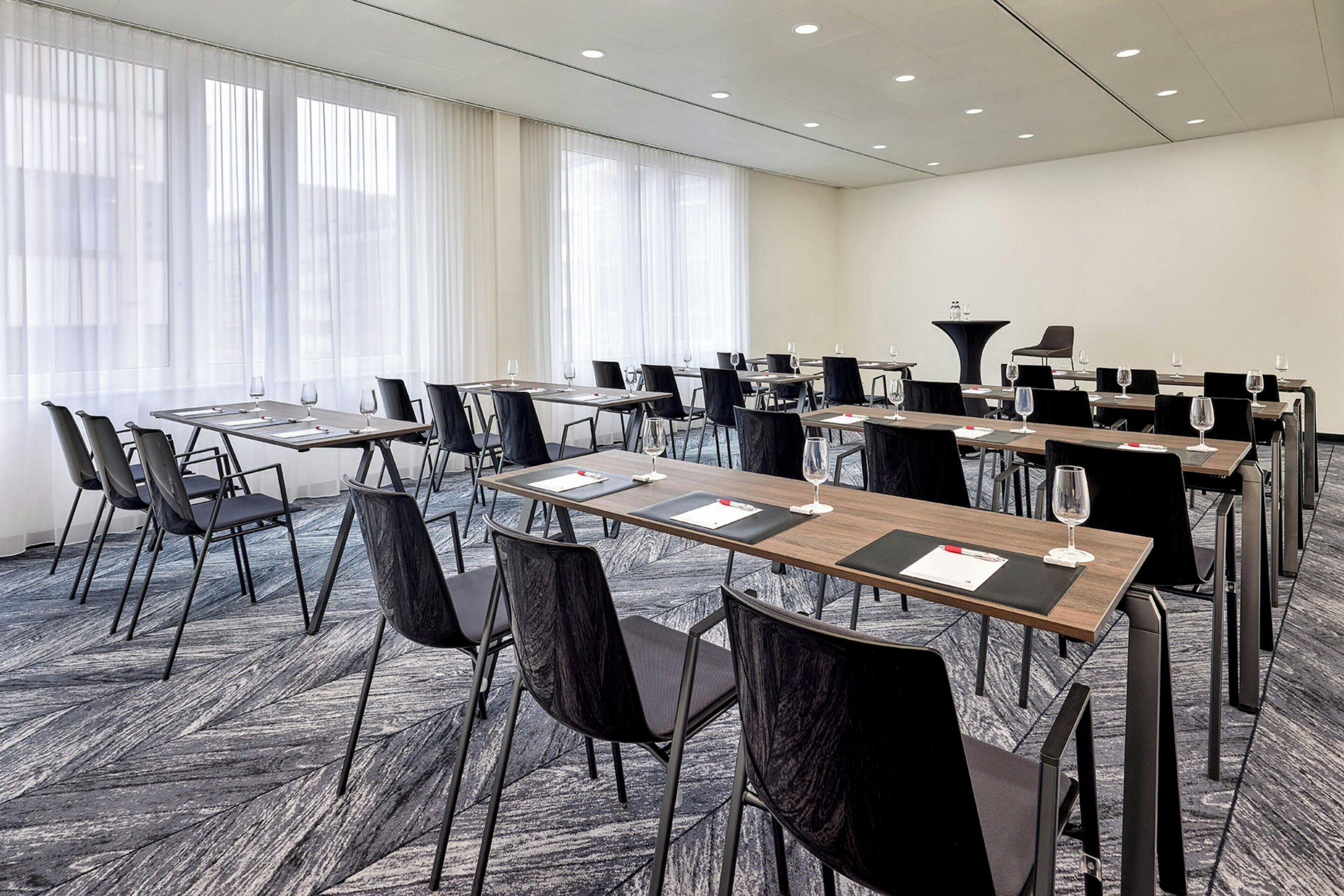A conference room with multiple tables and chairs, large windows with sheer curtains, and a carpeted floor.