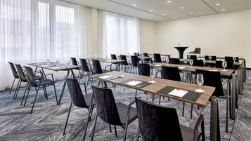 A conference room with multiple tables and chairs, large windows with sheer curtains, and a carpeted floor.