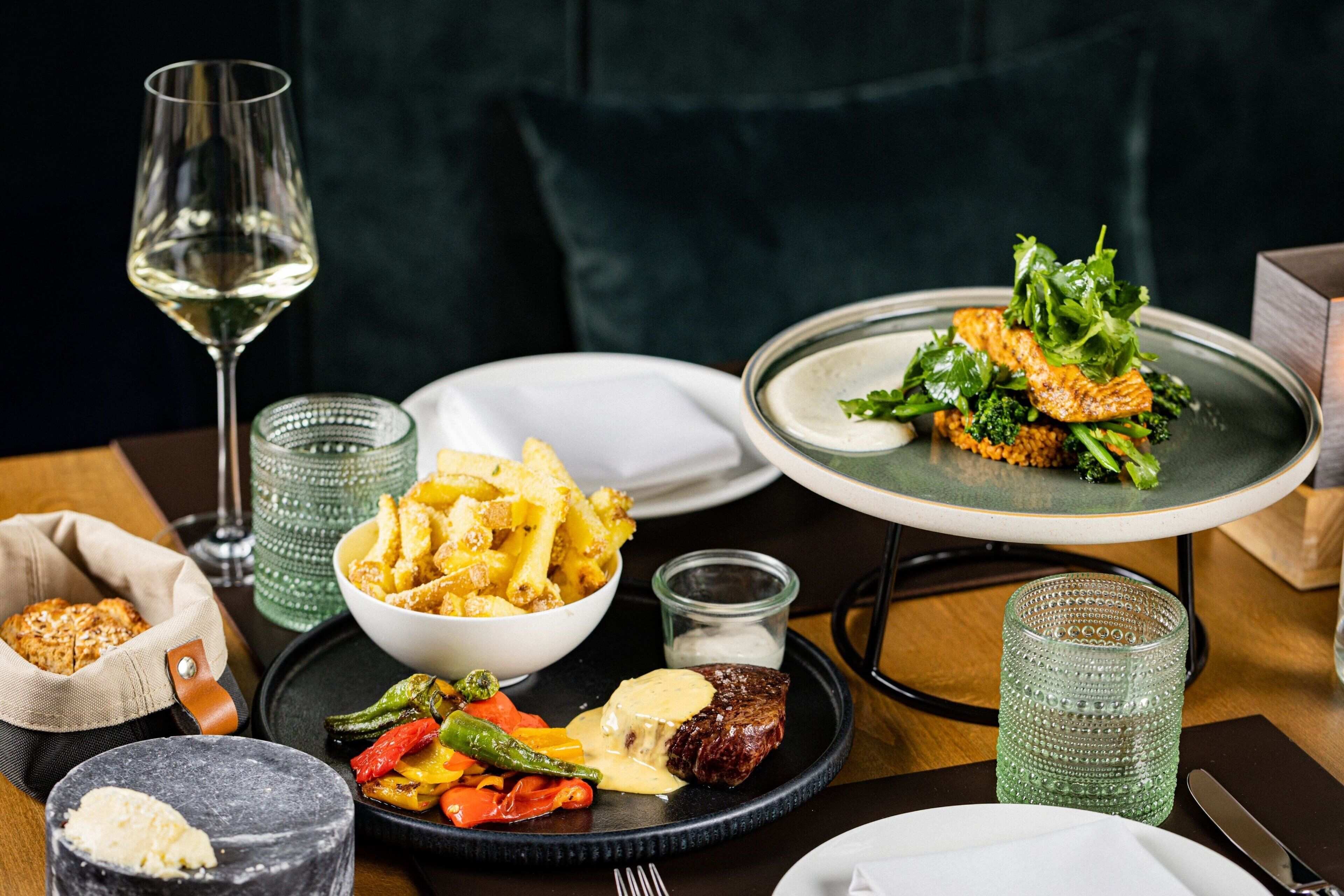 A dining table with various dishes, including a steak with sauce, vegetables, and a bowl of pasta. There are also glasses and a wine glass on the table.