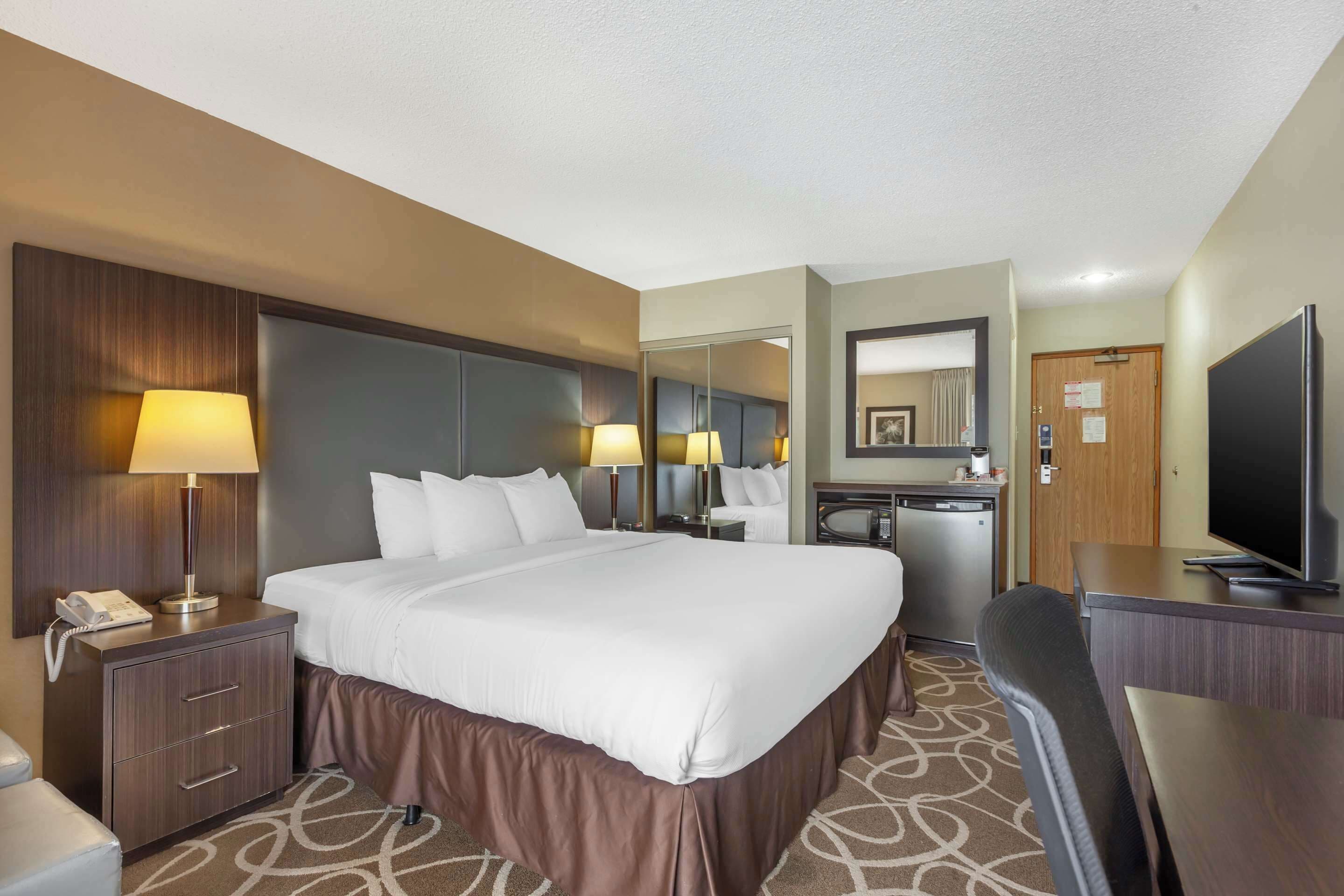 A hotel room with a large bed, bedside lamps, a desk with a television, a mirror, and a bathroom with a sink and toilet.