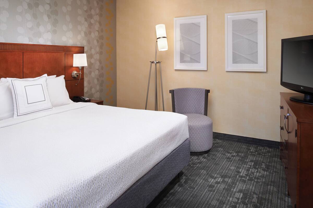 A hotel room with a large bed, a desk with a lamp, a chair, a television, and artwork on the walls.
