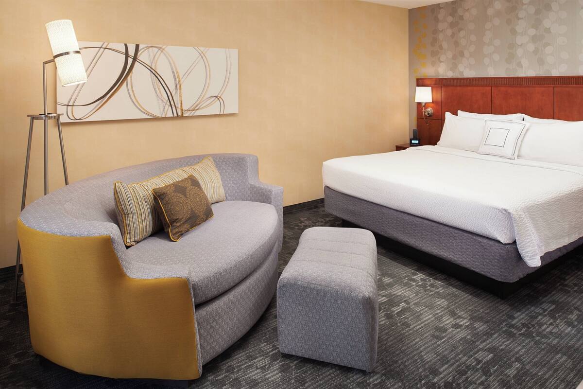 A hotel room with a curved sofa, a matching ottoman, a large bed, and a floor lamp.