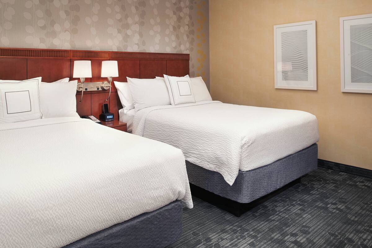 Een hotelkamer met twee bedden, een houten hoofdeinde, twee bedlampjes en twee ingelijste kunstwerken aan de muur.