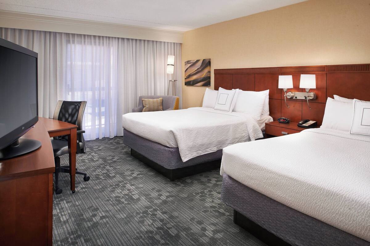 A hotel room with two beds, a desk, a TV, and a large window with curtains.
