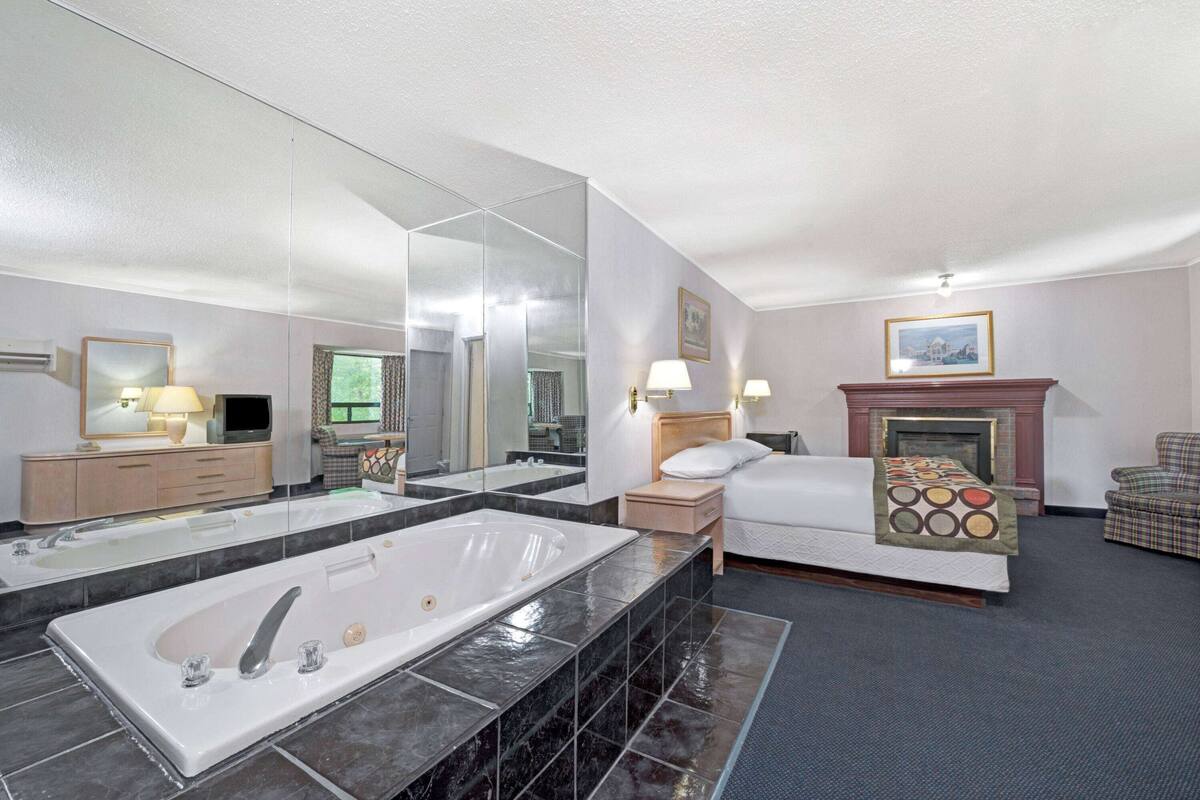 A modern hotel room with a large bathtub, a separate shower area, a bed with a headboard, a fireplace, and a seating area with a chair.