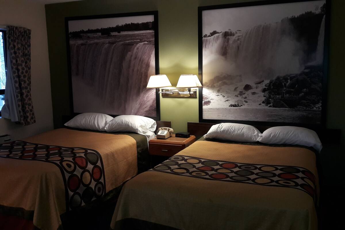 A hotel room with two beds, a nightstand with a lamp, a window with curtains, and two large waterfall paintings on the wall.