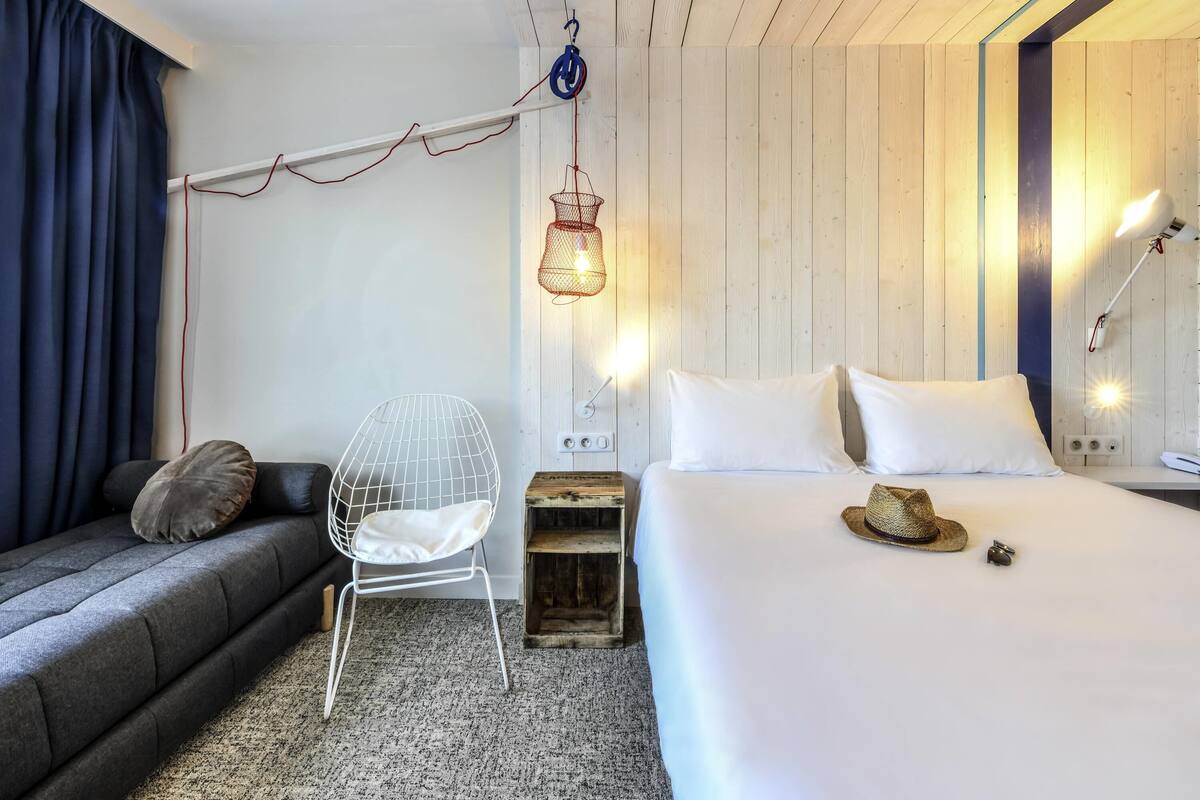 A modern hotel room with a grey sofa, a white wire chair, a bed with white linens, a woven hat, and a hanging light fixture.