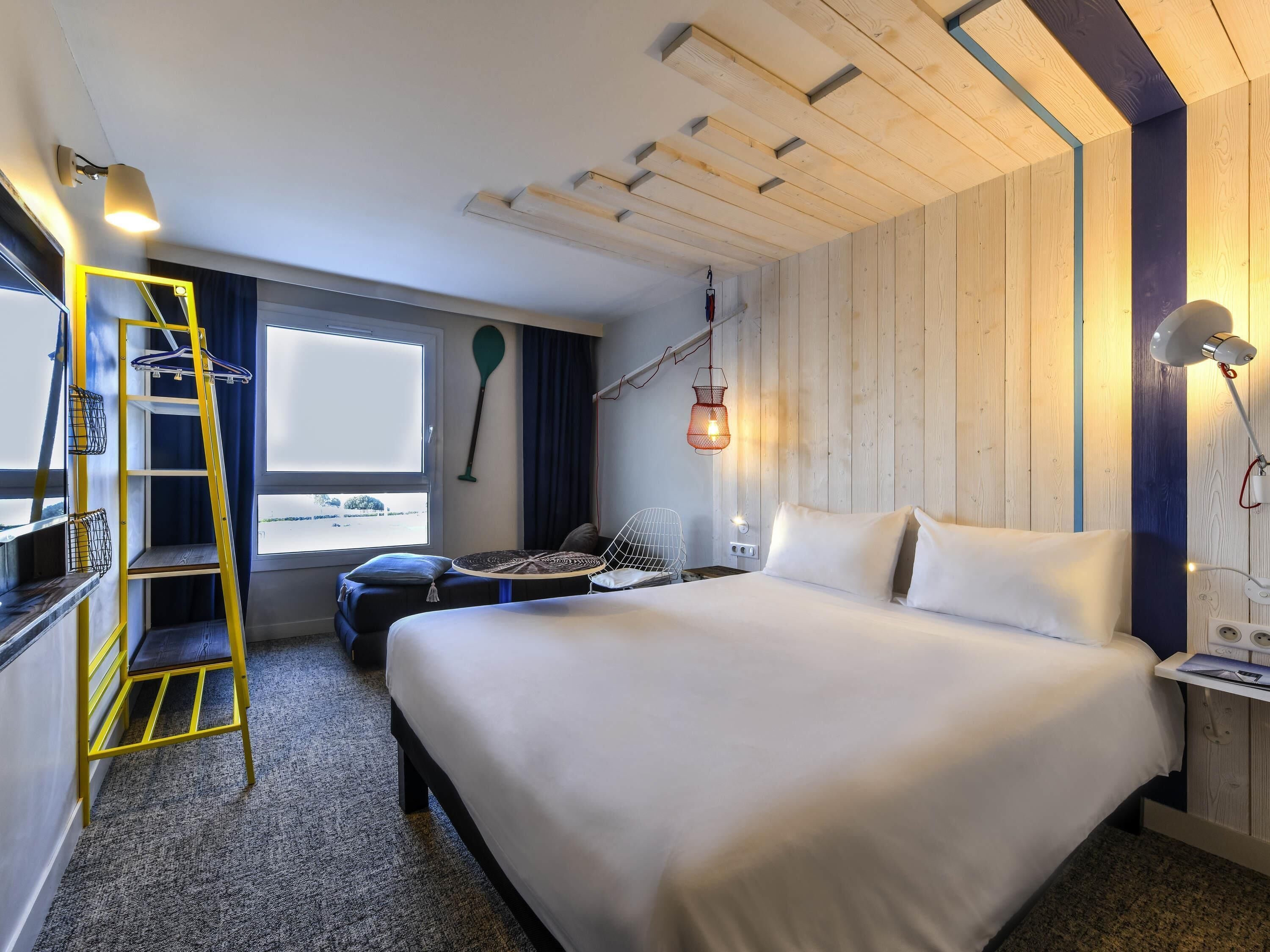 A hotel room with a bed, a desk with a chair, a small table, a ladder, a window, and a wall-mounted lamp.