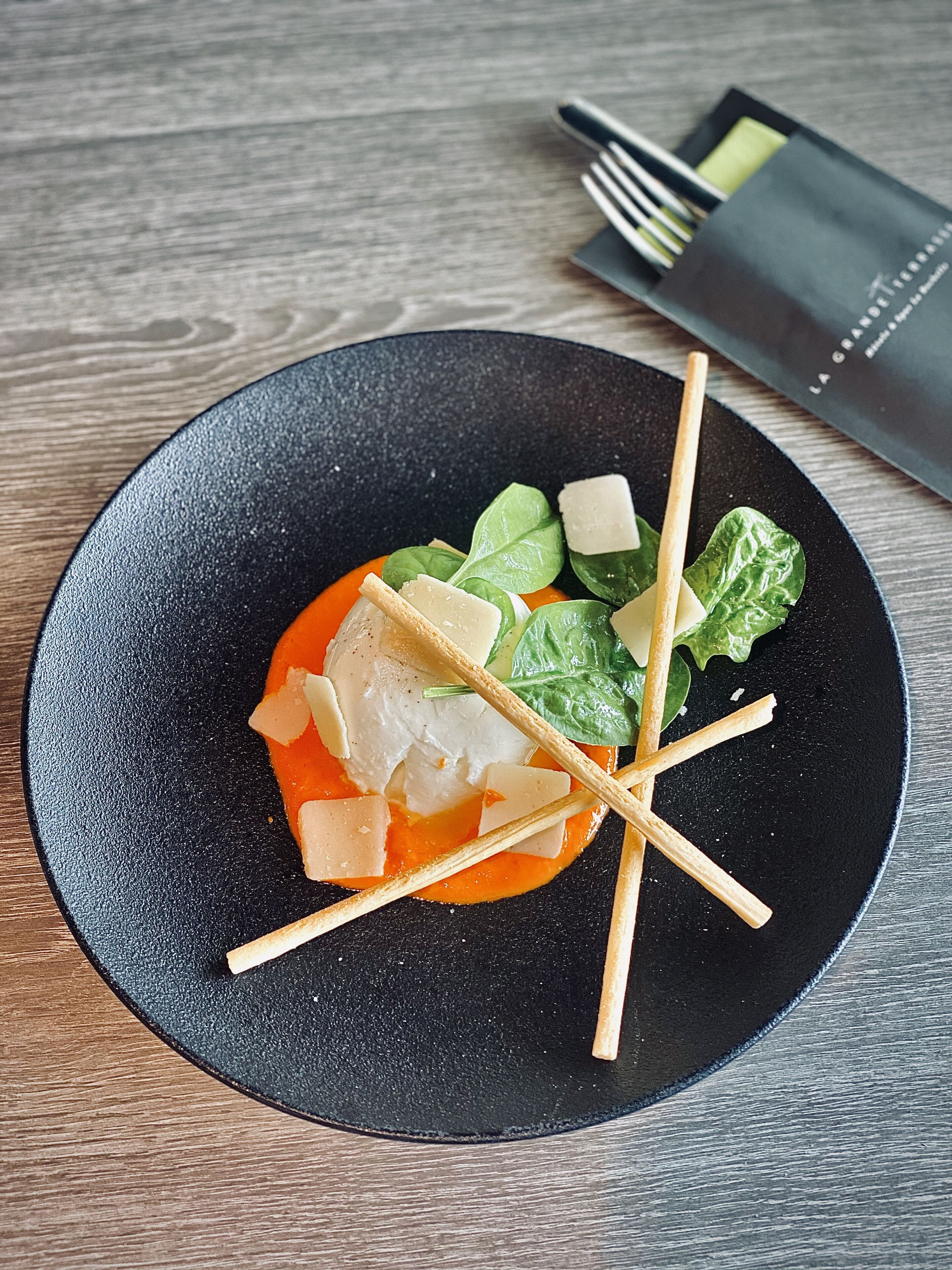 A black plate with a dish containing fish, cheese, and greens, accompanied by chopsticks and a napkin on a wooden table.