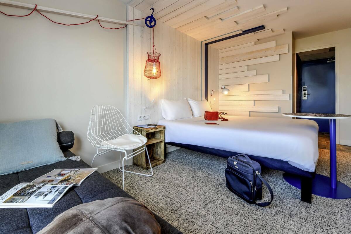 A hotel room with a bed, a small table, a chair, a desk, and a hanging light fixture.