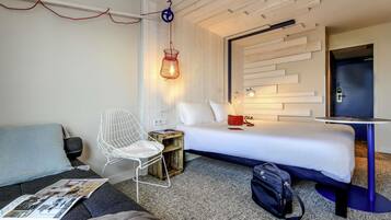 A hotel room with a bed, a small table, a chair, a desk, and a hanging light fixture.