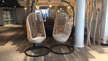 Two wicker hanging chairs with black frames in a modern interior space with wooden flooring and a bar area in the background.