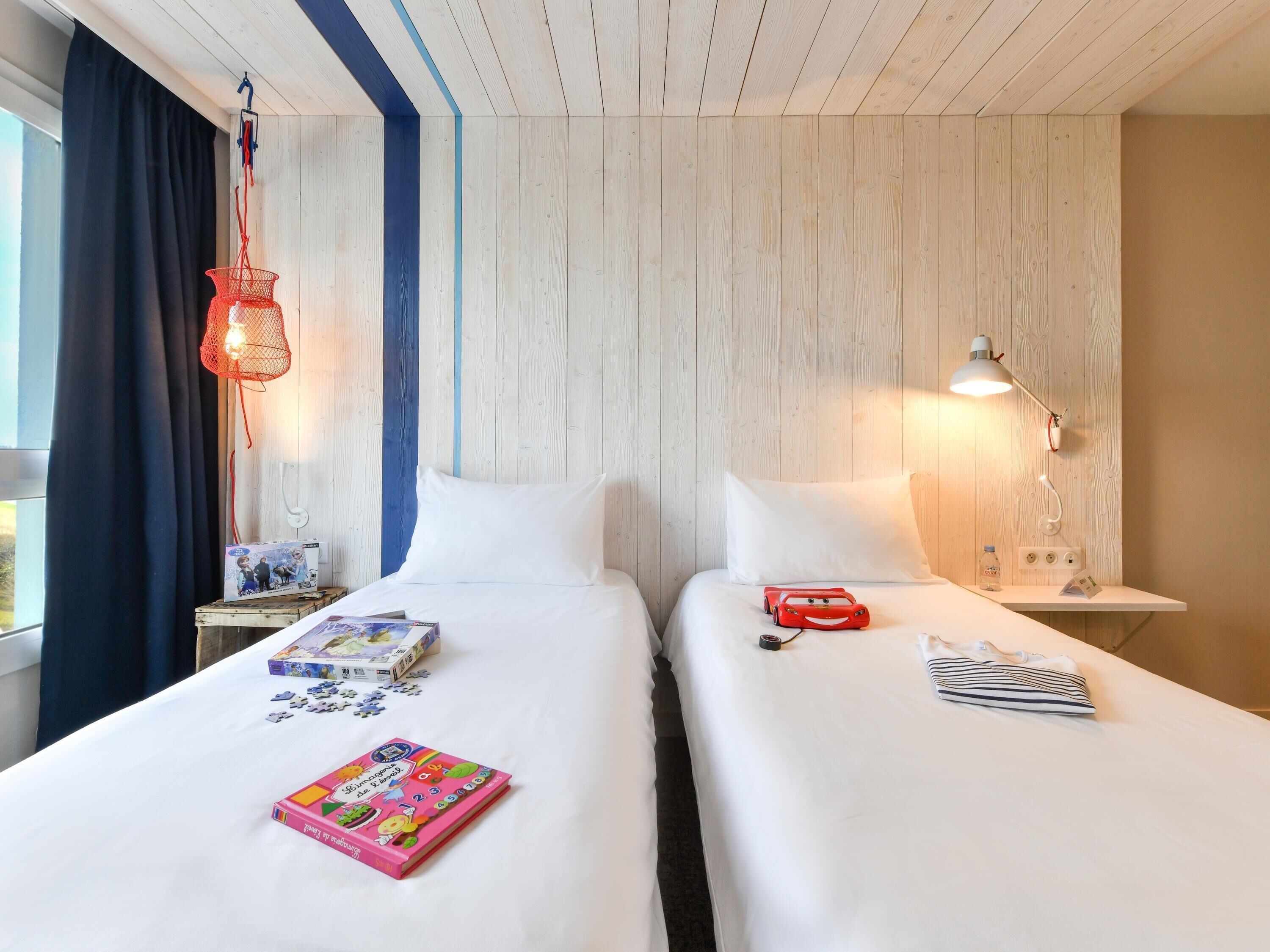 A hotel room with two beds, a window with curtains, a desk with a lamp, and a small table with a book and a toy car.