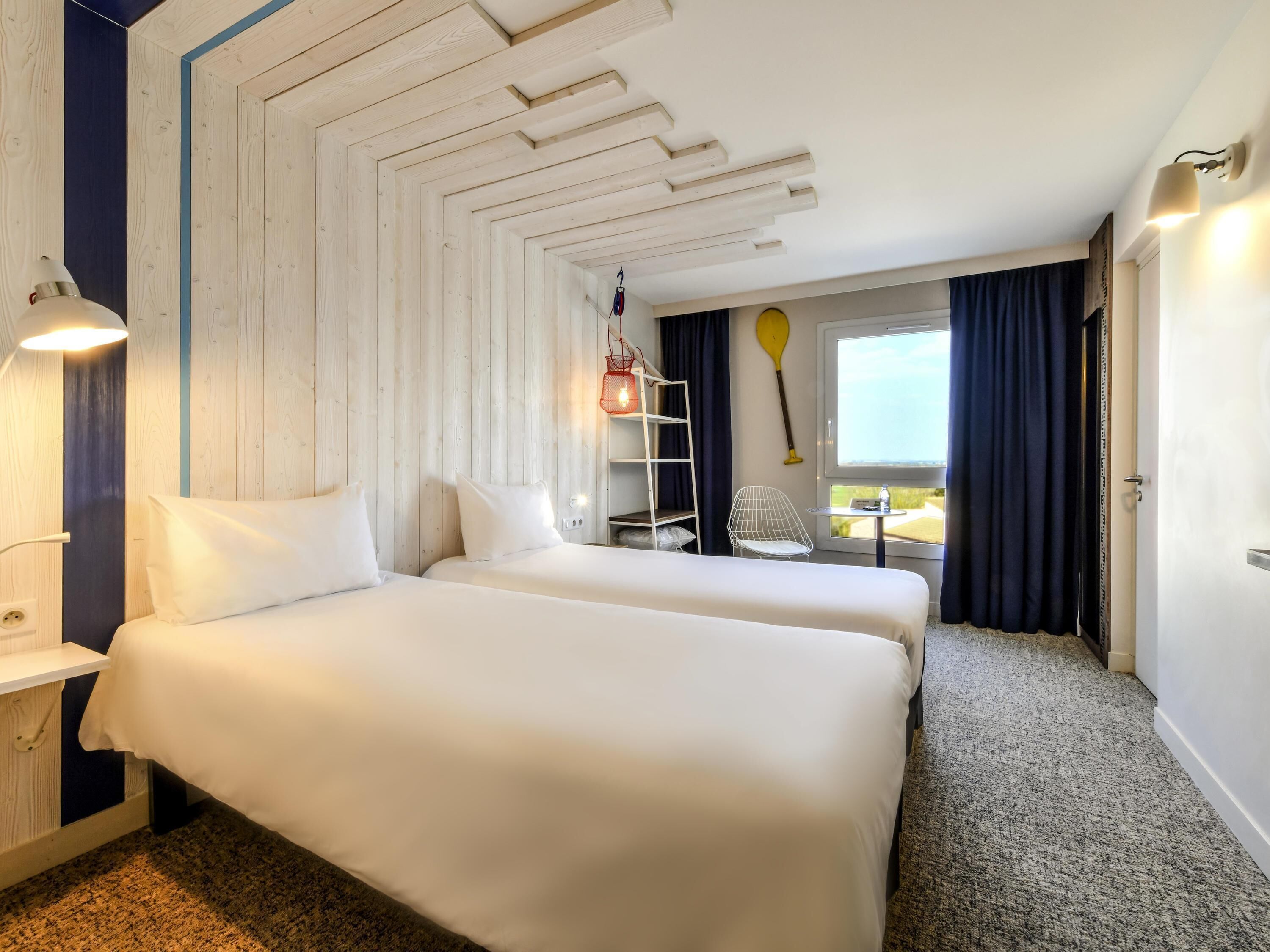 A hotel room with two beds, a desk, a chair, a window with curtains, and a view of the outdoors.