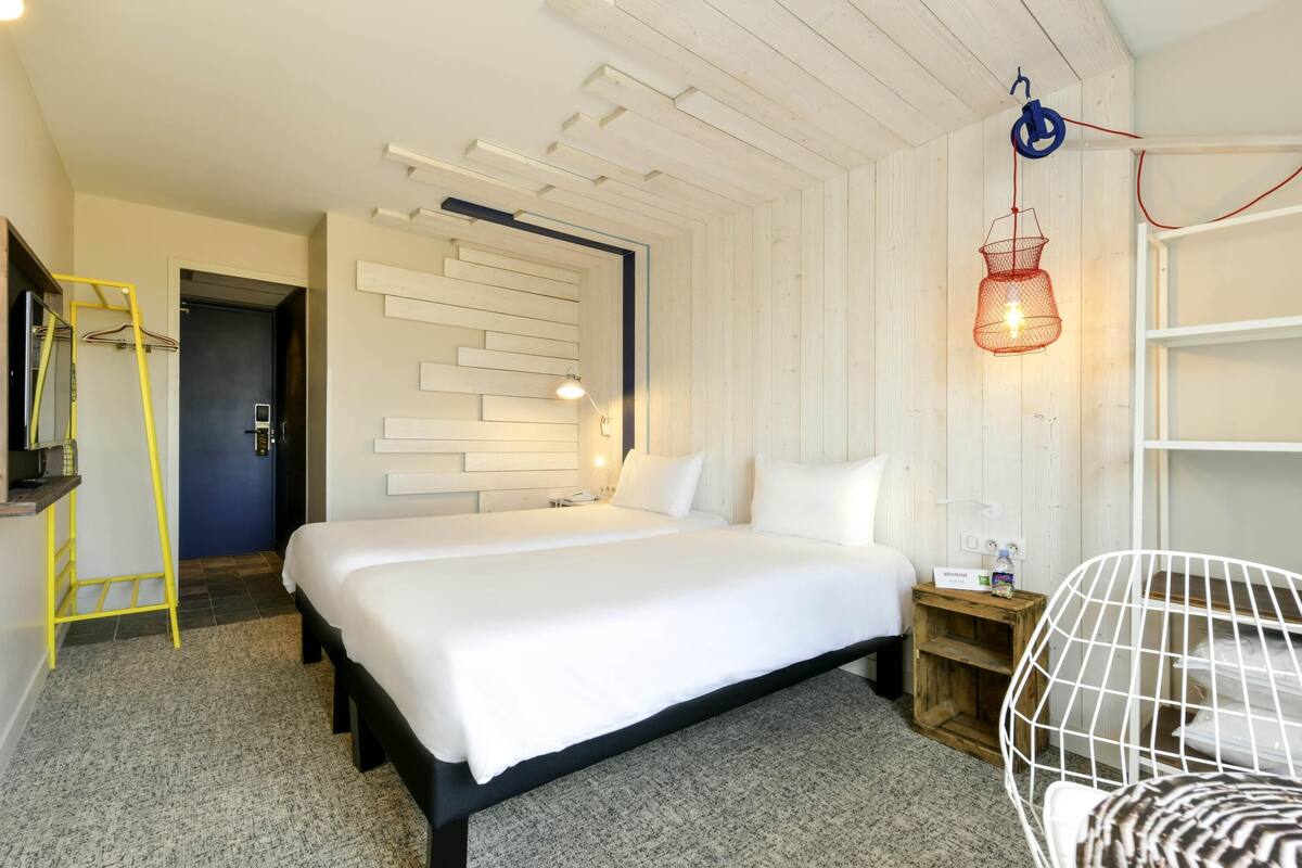 A hotel room with a bed, a desk, a chair, a ladder, a wall-mounted shelf, and a hanging light fixture.
